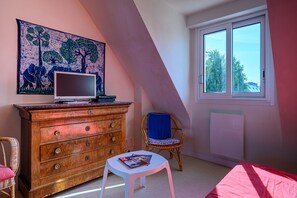 Un dormitorio con cómoda, televisor encima, una silla, una mesita y ventana con vista a árboles.