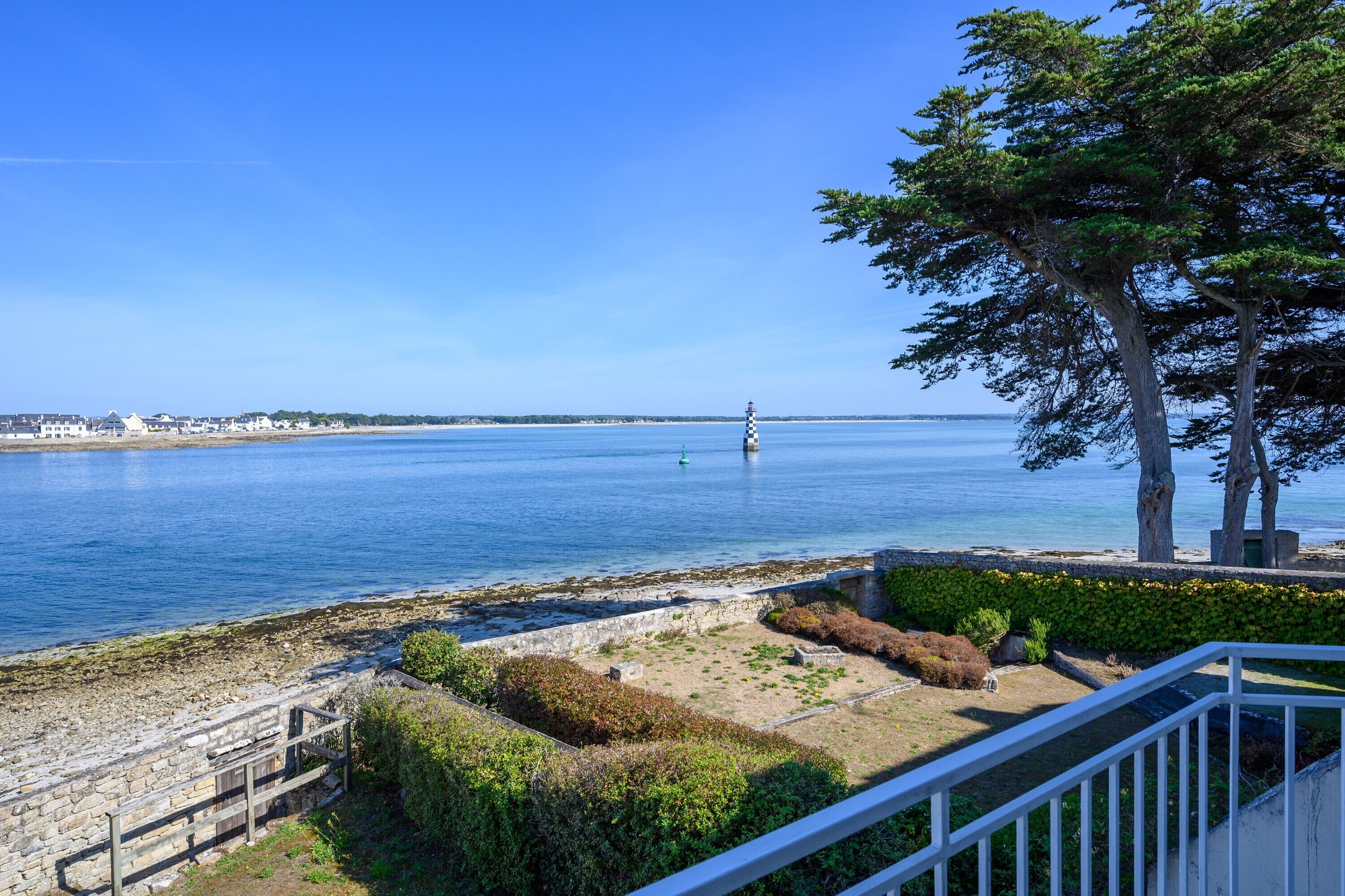 A coastal view with a lighthouse, stone wall, and a grassy area with bushes.