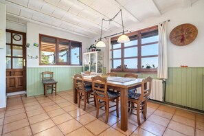 Un comedor con mesa y sillas de madera, un aparador con cristalería y ventanas amplias con cortinas.