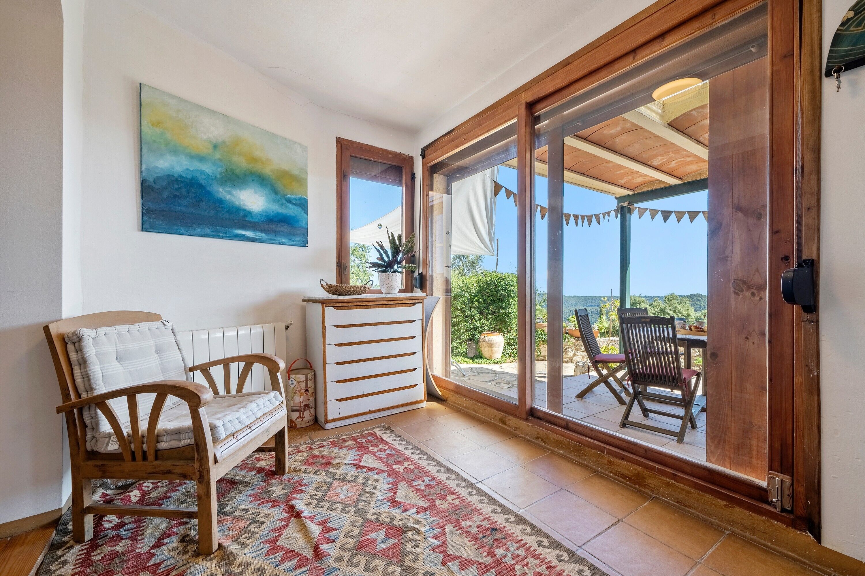 A room with a wooden bench, a white chest of drawers, a patterned rug, and a large window with a view of the outdoors.