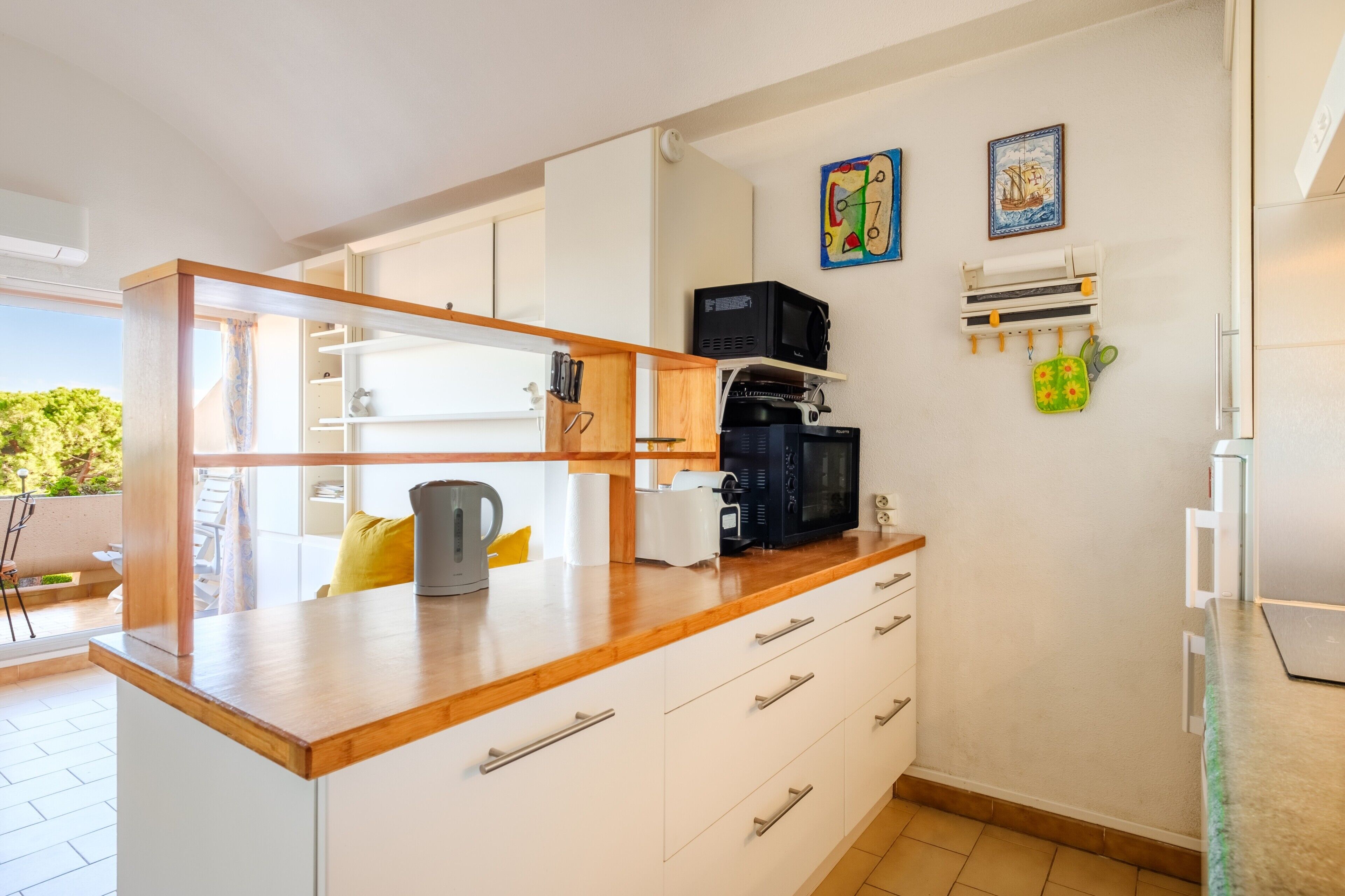 Une cuisine avec des meubles blancs, un plan de travail en bois, une bouilloire, un four à micro-ondes et une petite étagère contenant des ustensiles de cuisine.