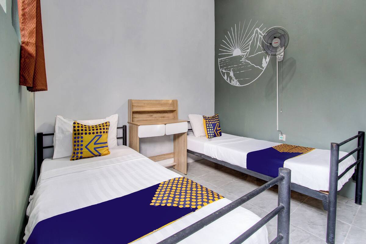 Una habitación con dos camas, un cabecero de madera, un ventilador de pared y un cuadro con un sol y un paisaje.