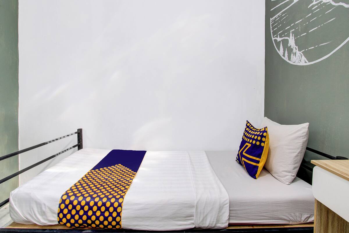 Una cama individual con una colcha blanca y azul a rayas, un almohadón con estampado azul y amarillo, y un mural en la pared que muestra un sol y un paisaje.