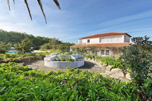 A white building with a red-tiled roof, surrounded by lush greenery and a circular concrete structure.