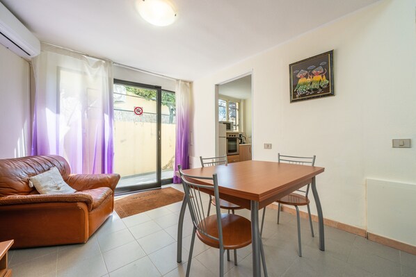 A living room with a brown leather sofa, a wooden dining table with four chairs, a kitchen area visible through an open door, and a framed picture on the wall.