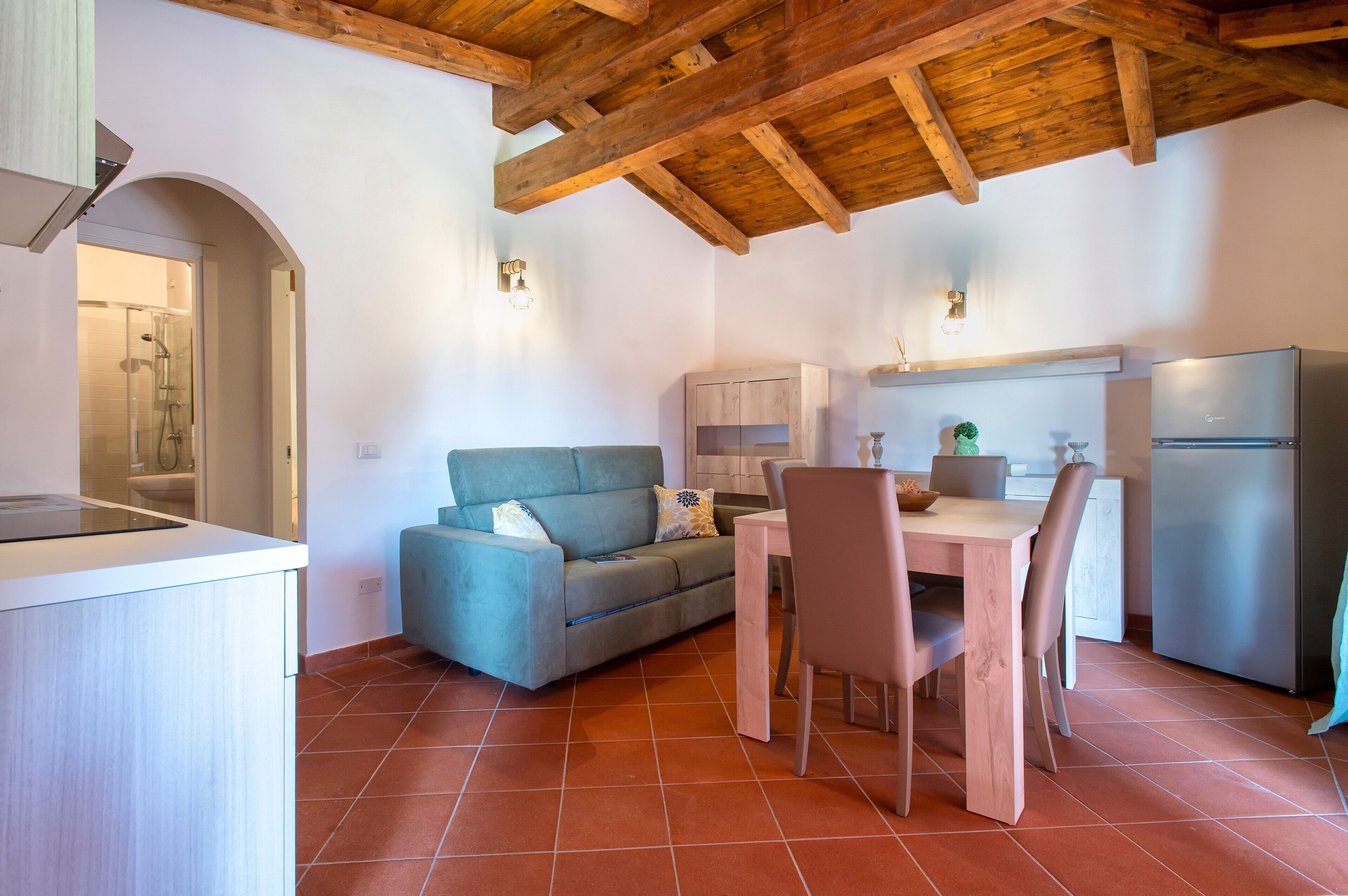 A compact kitchen with a wooden ceiling, a sofa, a dining table with chairs, and a refrigerator.