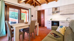 A kitchen with wooden beams, a dining area with a table and chairs, and a sofa with a magazine. - Vacation Home "Villa Robi" near Beach with Private Terrace & Shared Garden (stintino)