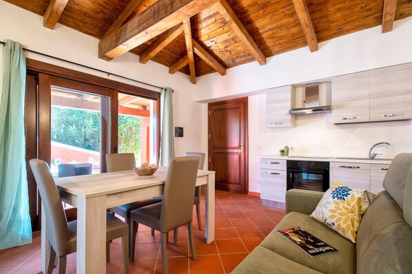 A kitchen with wooden beams, a dining area with a table and chairs, and a sofa with a magazine. - Vacation Home "Villa Robi" near Beach with Private Terrace & Shared Garden (stintino)