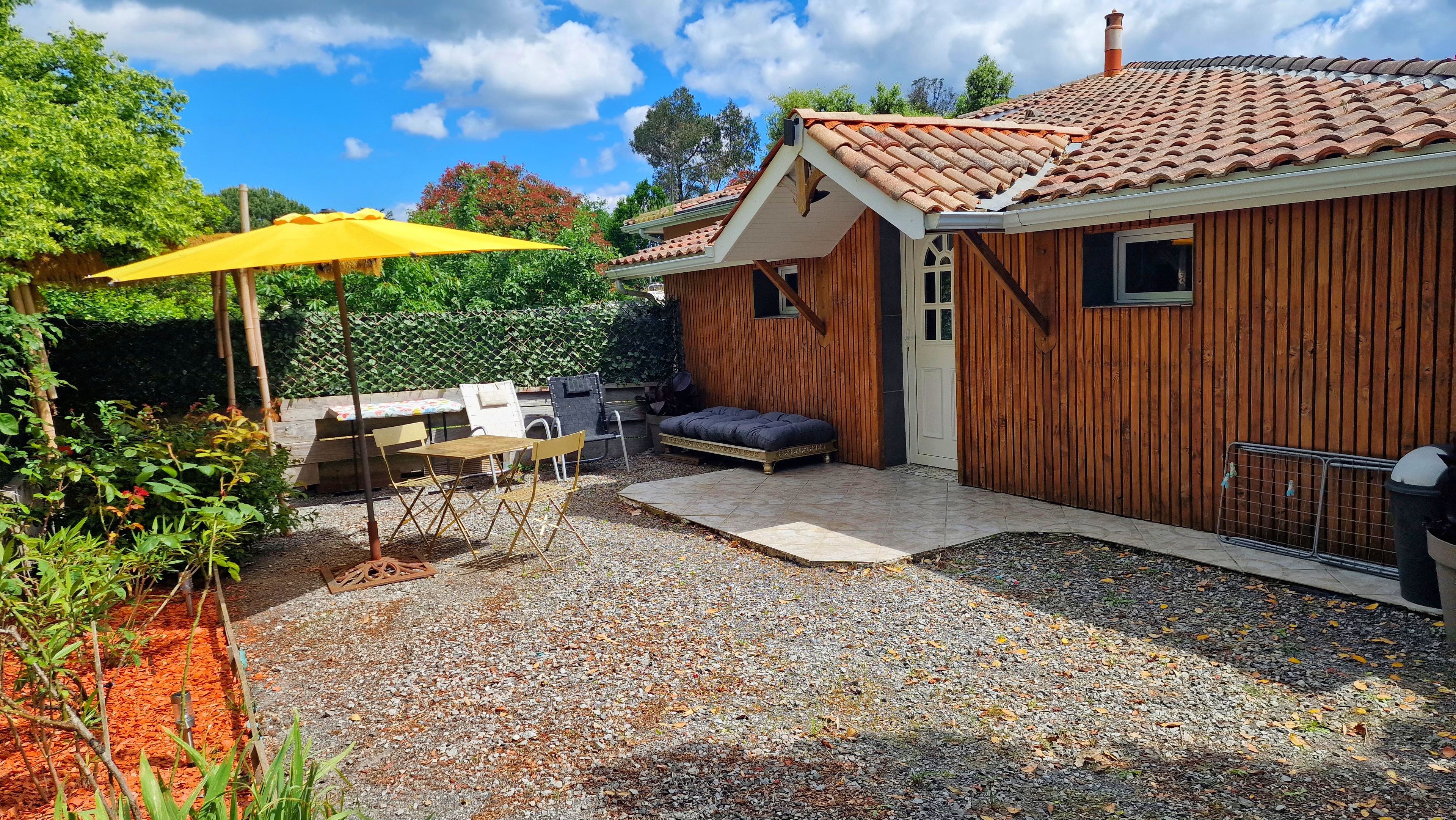 A wooden house with a tiled roof, a patio with a table and chairs, and a yellow umbrella.