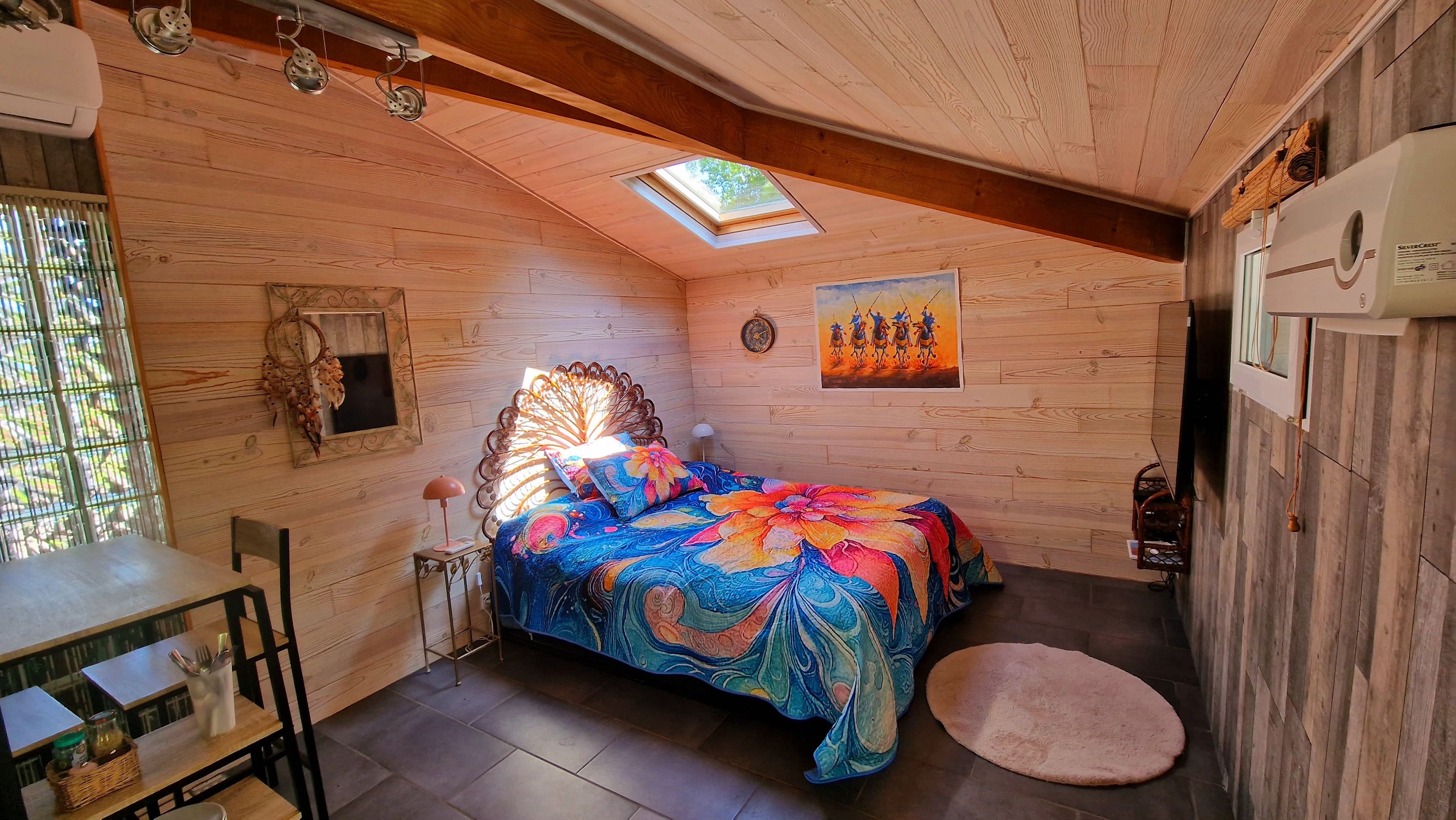 Een gezellige kamer met een bed, een klein tafeltje en een stoel. Boven het bed is een dakraam en aan de muur hangt een schilderij.