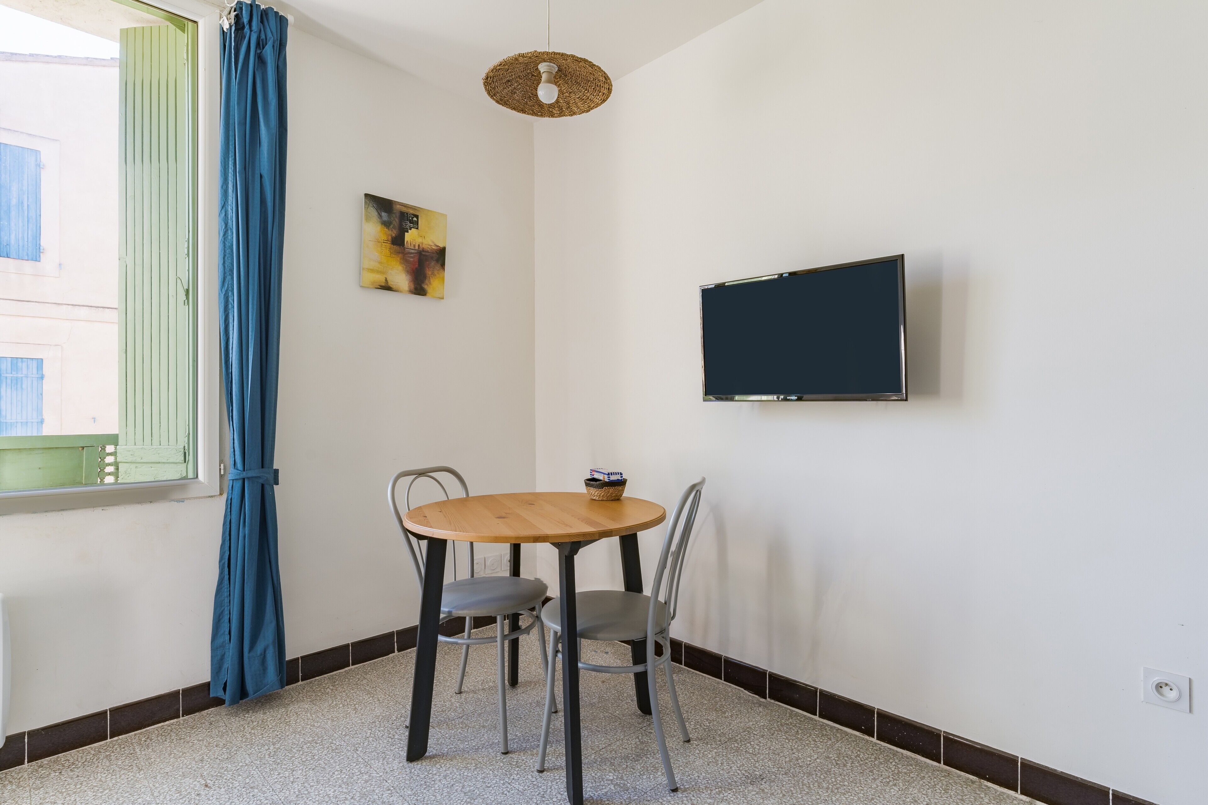 Ein Zimmer mit einem Holztisch, zwei Stühlen, einem Fernseher an der Wand, einem Fenster mit blauen Vorhängen und einem Bild an der Wand.