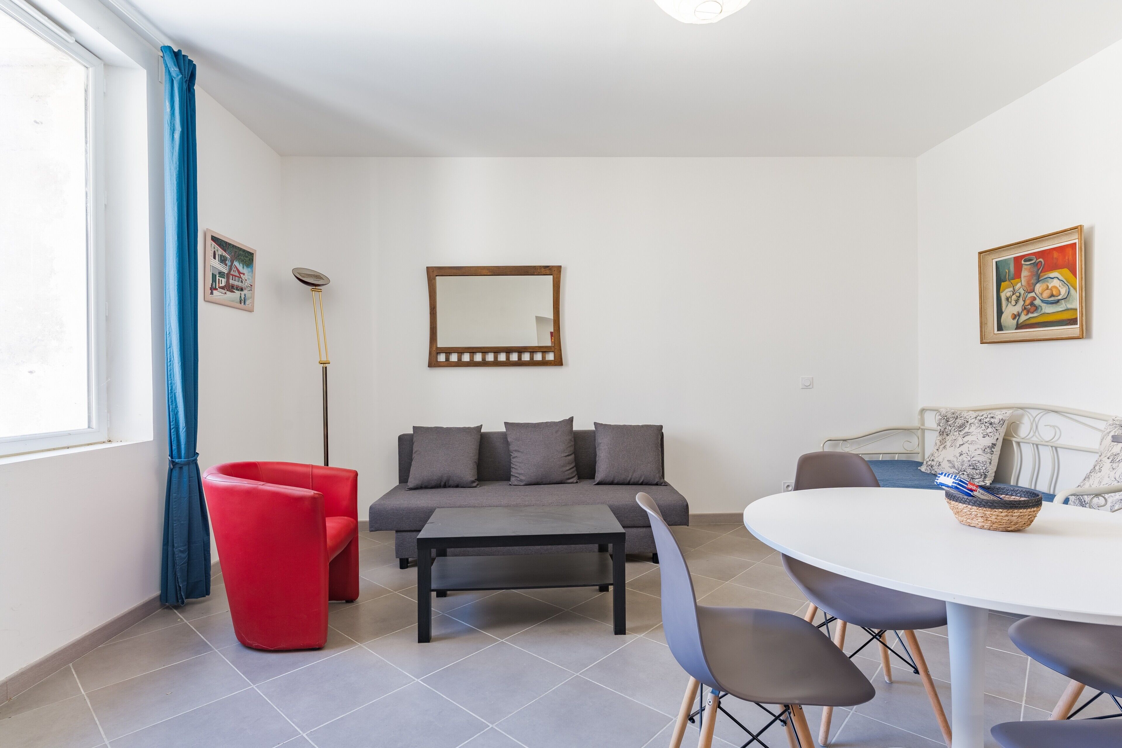 A room with a grey sofa, red chair, round dining table, and a mirror on the wall.