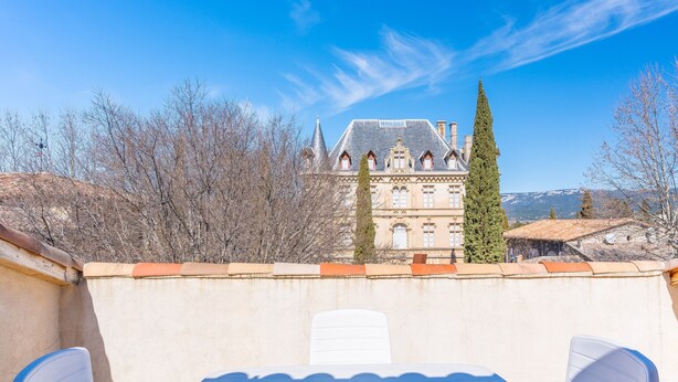 Terraço com mesa e cadeiras, com vista para um edifício histórico com telhado inclinado e uma árvore alta.