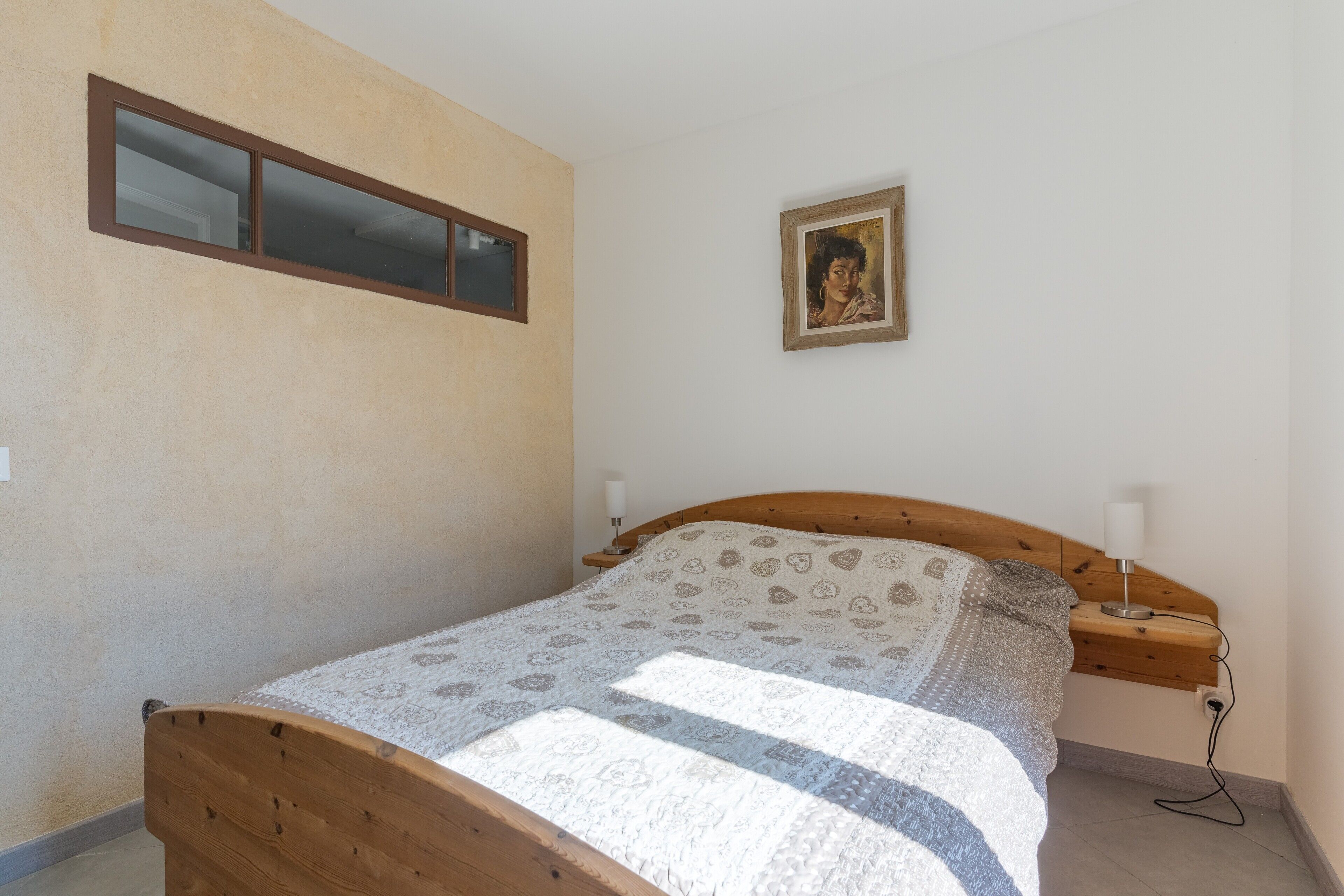 A bedroom with a wooden bed, a bedside table with a lamp, a wall-mounted picture, and a window with blinds.