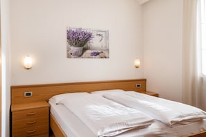 Un dormitorio con cabecera de madera, cama con ropa de cama blanca, mesita de noche con cajones, lámpara de pared y un cuadro con flores de lavanda.