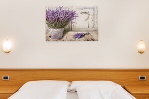 Un lit avec du linge de lit blanc, un tableau représentant de la lavande accroché au mur et deux appliques murales.