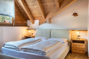 A bedroom with a wooden ceiling, a bed with white bedding, two bedside tables with lamps, and a window with a view of trees.