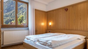 Quarto com cama de madeira, janela com vista para uma floresta, radiador e luminária fixada na parede.