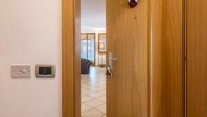 Entrada de um quarto de hotel com porta de madeira, parede branca com dois interruptores e vista para o interior do quarto, onde se vê uma cama e uma janela.