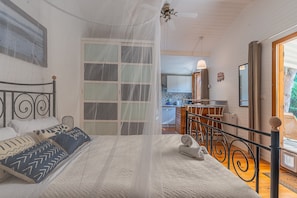 A bedroom with a bed, a window with curtains, a bedside table, and a view of the kitchen.