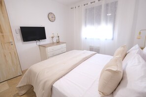 Une chambre à coucher avec un lit, un téléviseur fixé au mur, une commode en bois, une horloge et une fenêtre avec des rideaux légers.