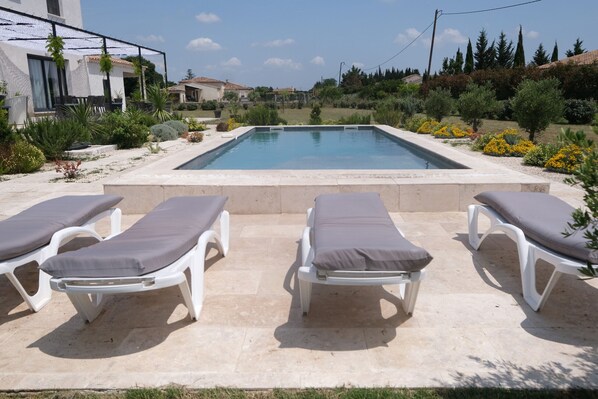 Une piscine entourée de quatre chaises longues, elle-même bordée d’un jardin aménagé avec différentes espèces de plantes, et offrant une vue sur les maisons situées au loin.