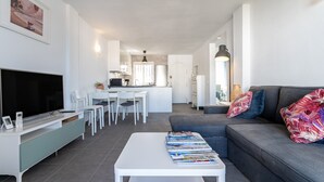 Un salon moderne avec un téléviseur à écran plat, une table basse blanche sur laquelle sont posés des magazines, un canapé gris agrémenté de coussins colorés, et une cuisine en arrière-plan.