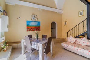 A dining area with a table and chairs, a sofa, a painting on the wall, and a staircase.