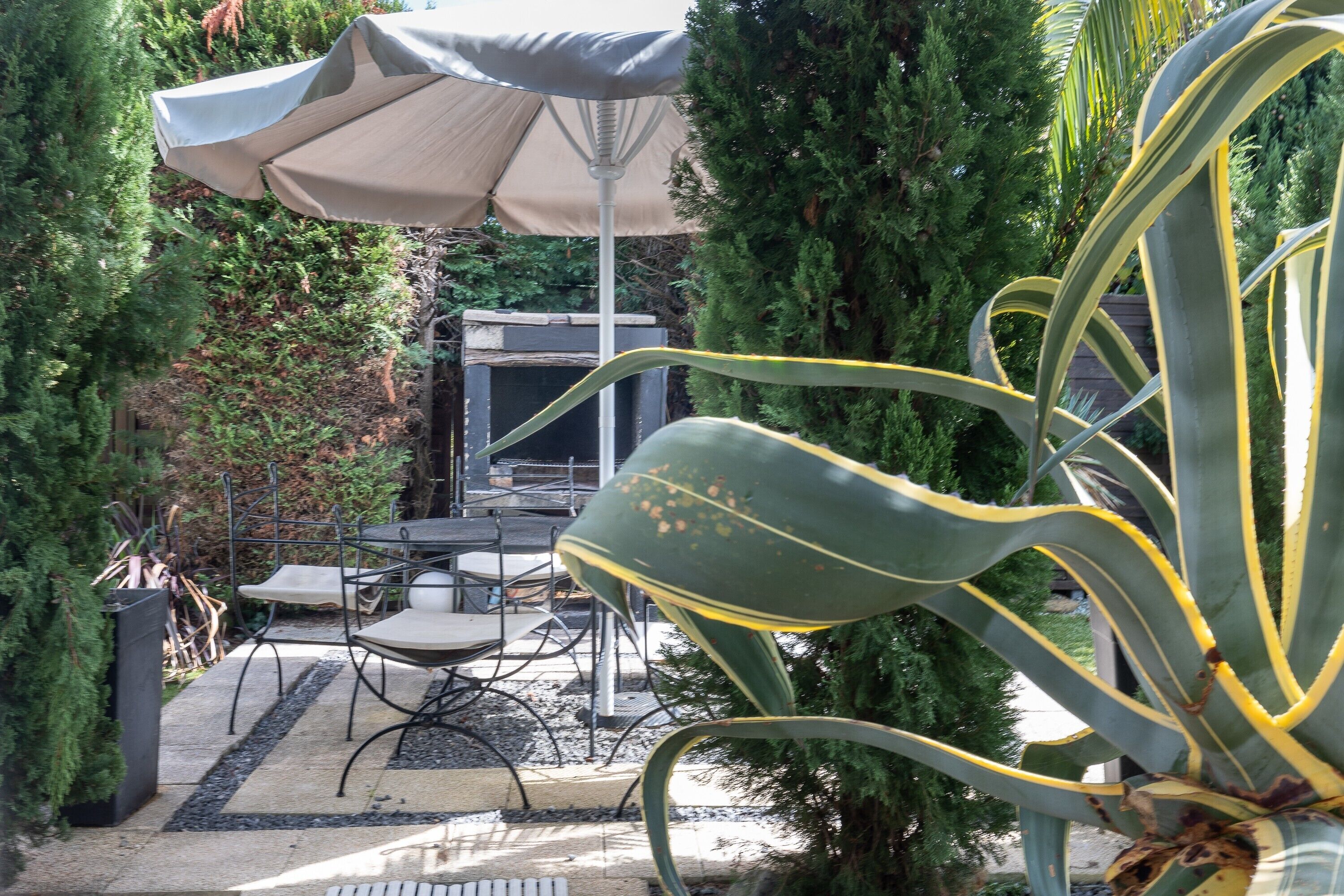 An outdoor patio with a table and chairs, surrounded by greenery and a large umbrella.