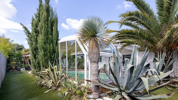 Une maison moderne dotée d’une véranda en verre, d’une piscine et de diverses plantes tropicales.