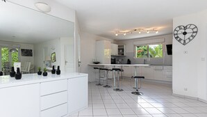 Une cuisine moderne avec des meubles blancs, un îlot central doté de tabourets de bar, une horloge en forme de cœur accrochée au mur et de grandes fenêtres.