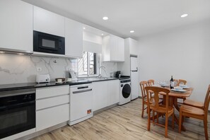 A modern kitchen with white cabinetry, a built-in microwave, a dishwasher, and a washing machine. There is a dining area with wooden chairs and a table set for two.