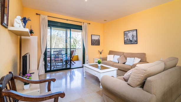 Una sala de estar con sofá, mesa de centro y una silla de madera. Hay un balcón con vistas a árboles.