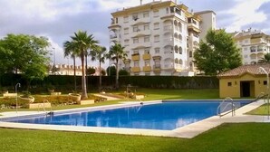Una piscina con césped, palmeras y un edificio de varios pisos al fondo.