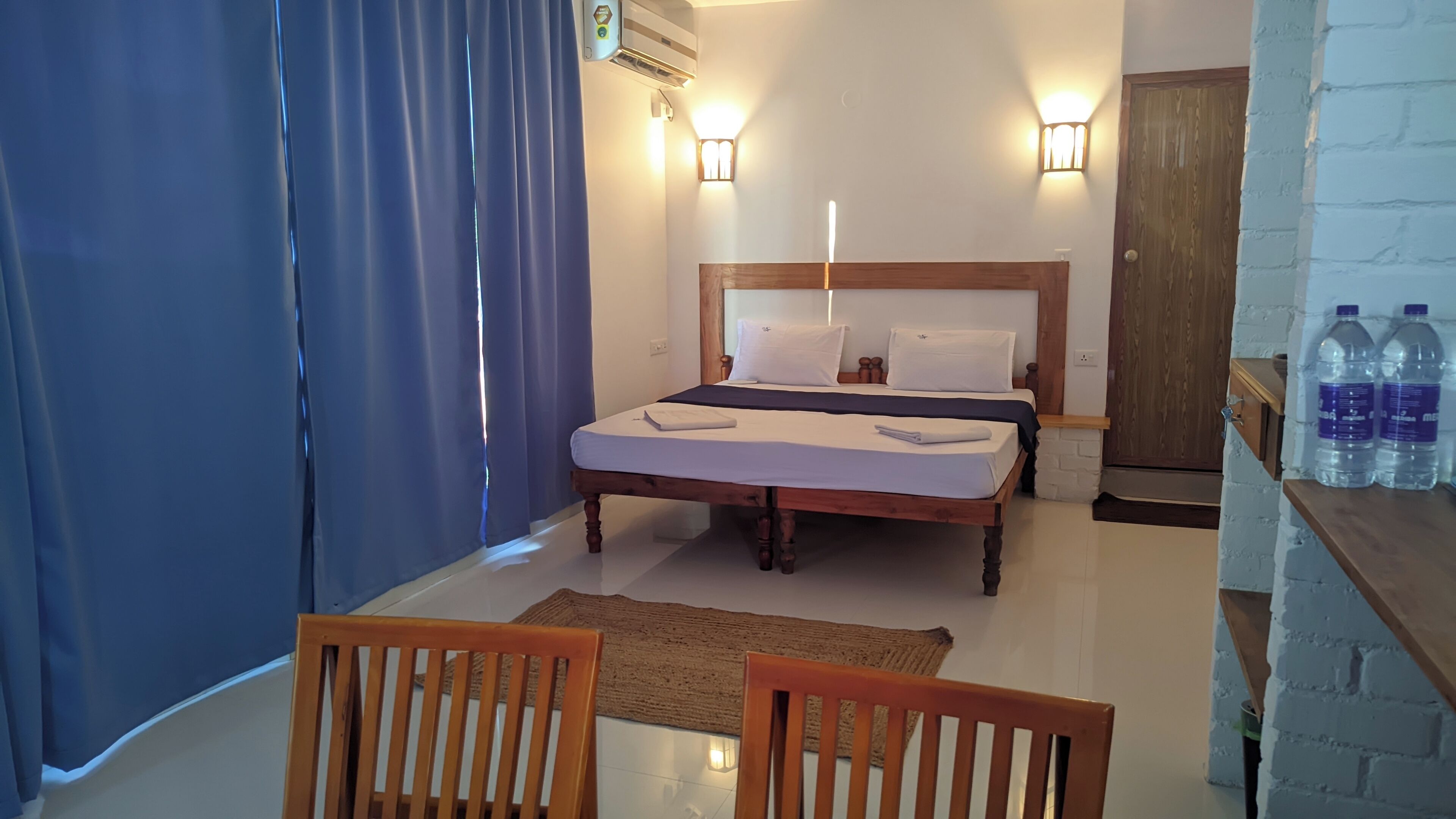 A hotel room with a bed, two wall-mounted lights, a wooden door, and a small table with water bottles.