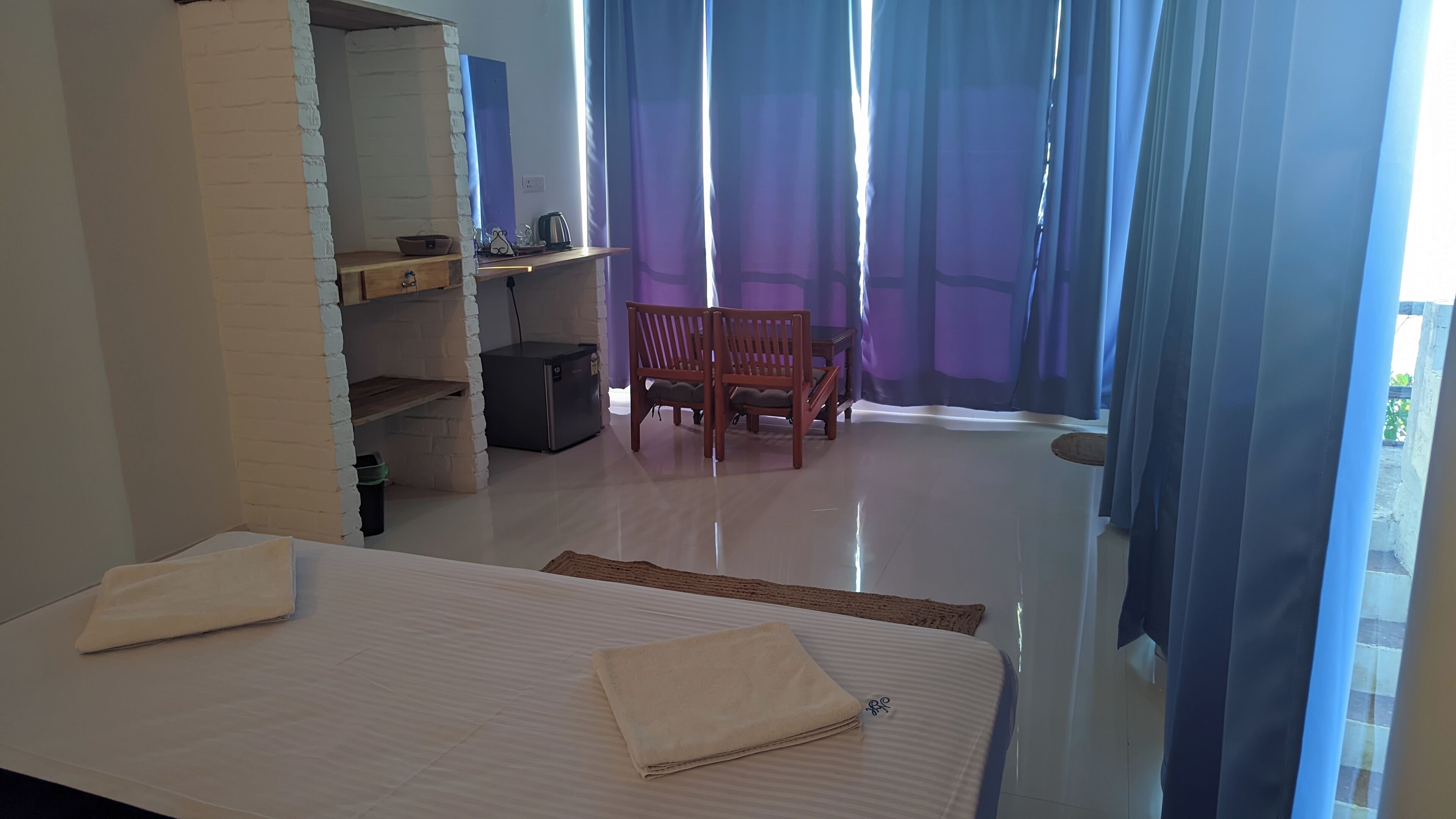 A hotel room with a bed, a wooden chair, a small table, and a blue curtain.