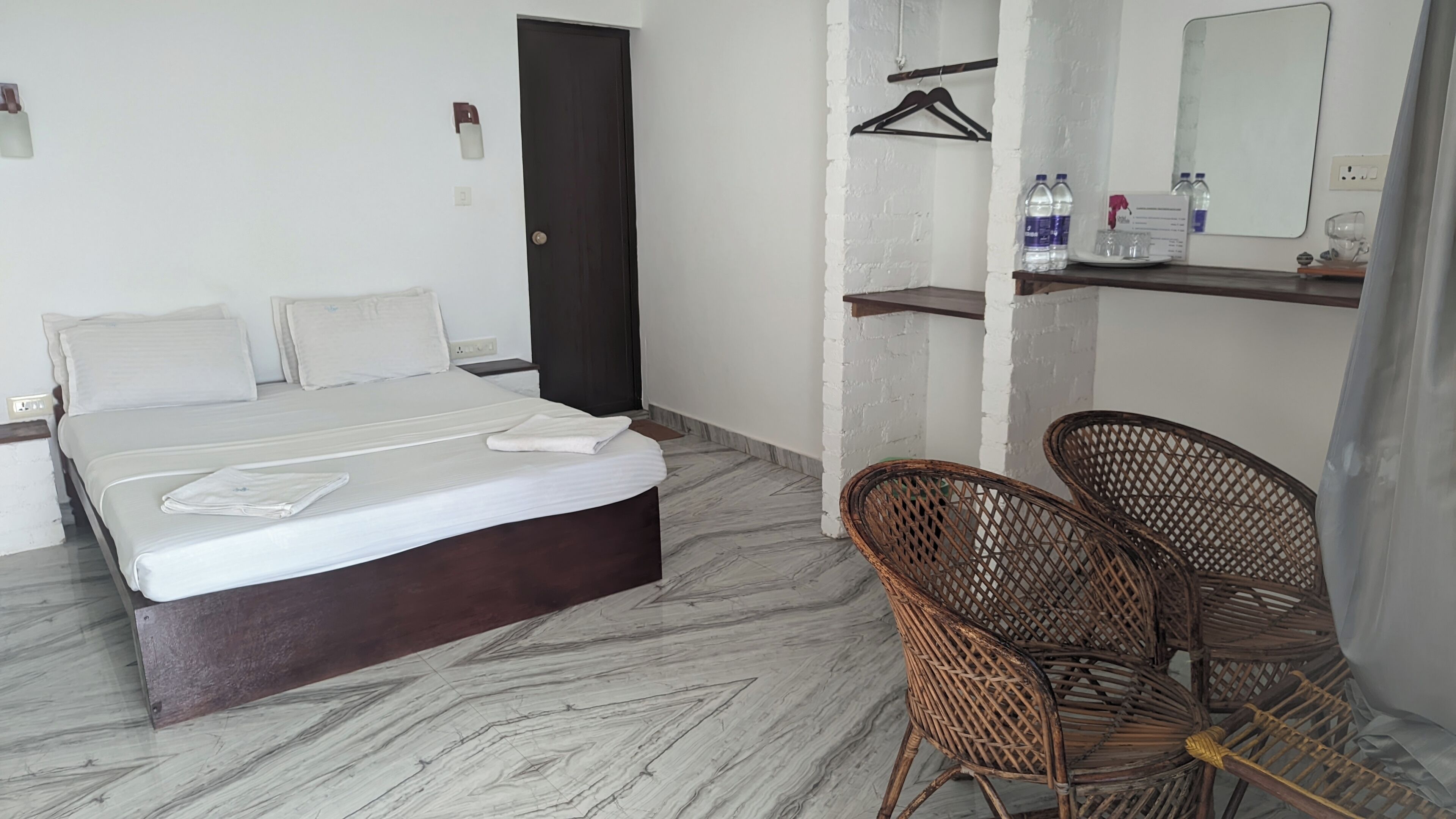 A hotel room with a bed, two chairs, a wardrobe, a mirror, and a shelf with toiletries.