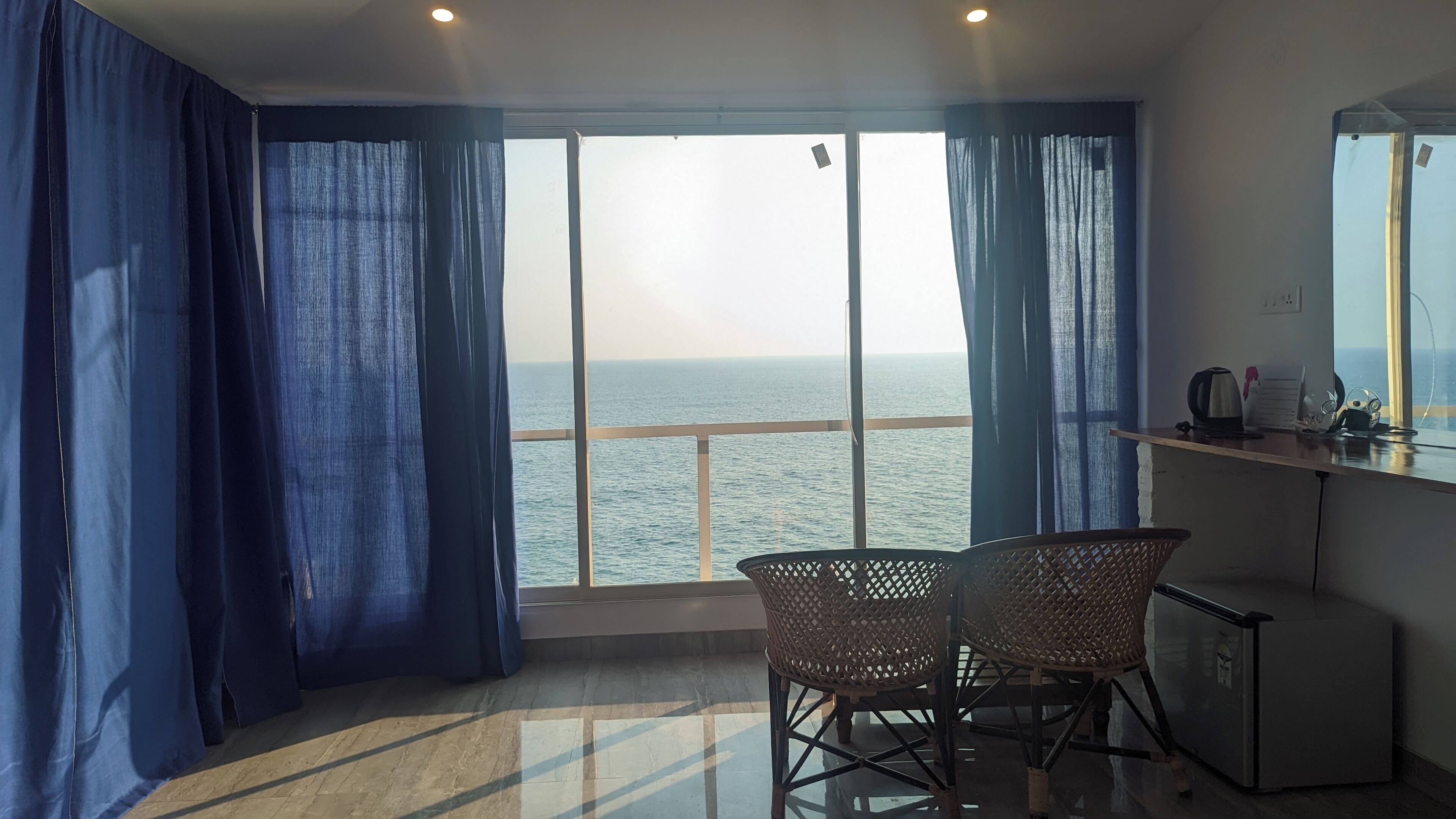A room with a large window overlooking the sea, a small table with chairs, and a kitchenette area.