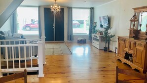 A spacious living room with a wooden floor, a white railing, a chandelier, and a large window with curtains.