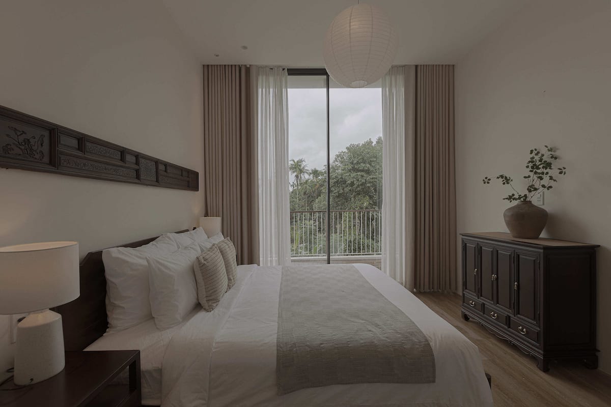 A hotel room with a large bed, a nightstand with a lamp, a dark wooden cabinet, and a view of greenery through the window.