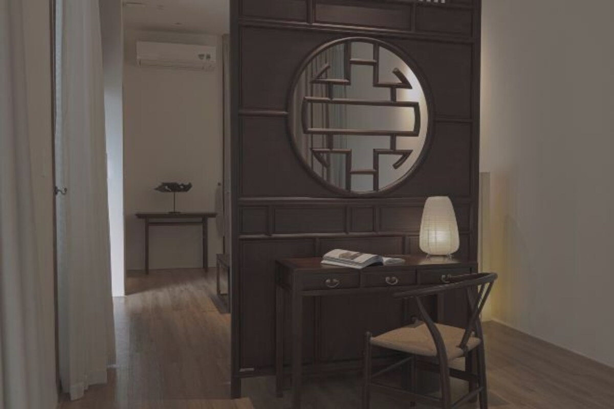Quarto com escrivaninha de madeira escura, uma cadeira, uma luminária e um painel decorativo com um espelho circular.
