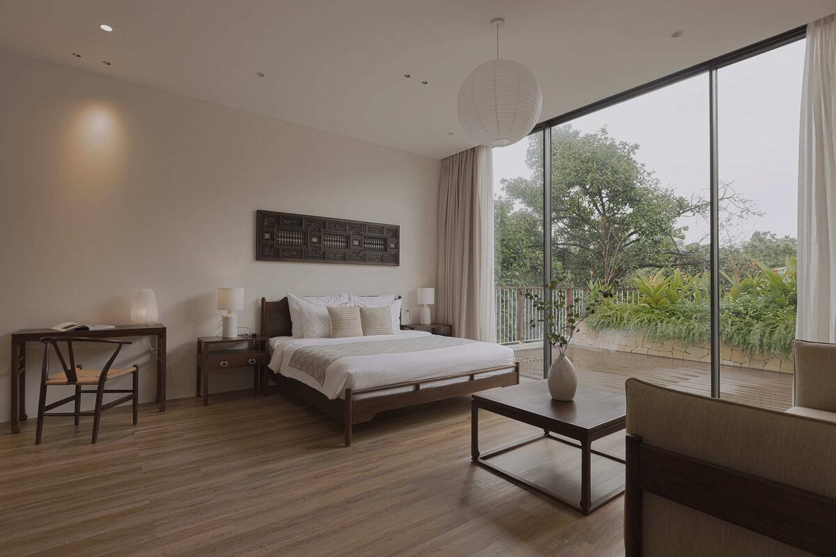 A modern bedroom with a large bed, a desk, a chair, and a view of greenery through a large window.
