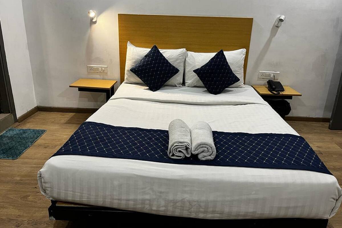 Una cama bien hecha con una colcha azul y blanca, dos almohadas y una toalla enrollada sobre la cama. Hay dos mesitas de noche, una de ellas con una lámpara y un teléfono.