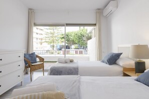 A hotel room with two beds, a wicker chair, a white dresser, and a view of the outside.