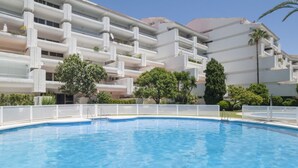 A swimming pool surrounded by a white fence, with a multi-story building featuring balconies and trees in the background. - Isabel Apartment - Golden Mile (Marbella)