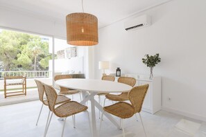 A modern dining area with a white table and wicker chairs, a hanging wicker light fixture, and a view of trees outside.