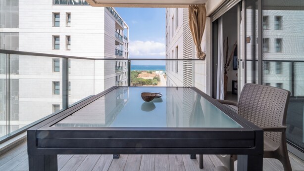 A balcony with a glass table and chair, overlooking a cityscape and the sea. - Beach Haven 3BR Apt by Sea N' Rent (Nahariya)