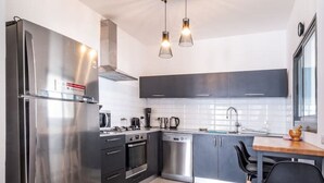 A modern kitchen with stainless steel appliances, a dishwasher, and a dining area with a wooden table and black chairs. - Beach Haven 3BR Apt by Sea N' Rent (Nahariya)