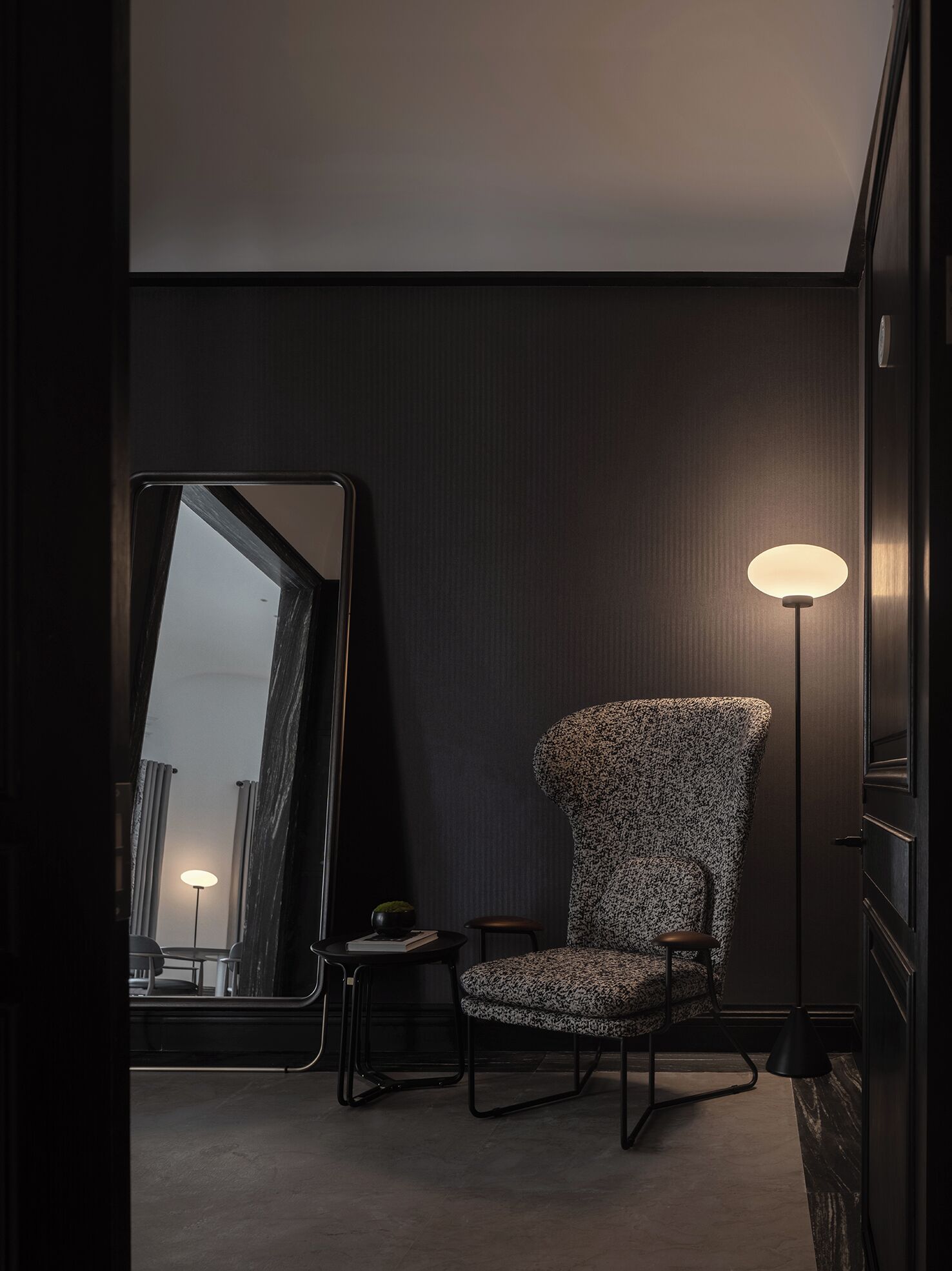 A modern hotel room with a large mirror, a patterned armchair, a small side table, and a floor lamp.
