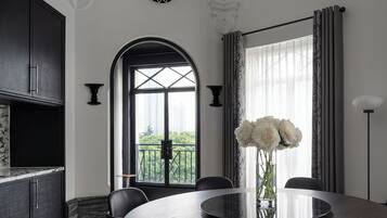 A modern dining room with a round table, black chairs, and a vase of white flowers. There is a large window with curtains and a view of greenery outside.